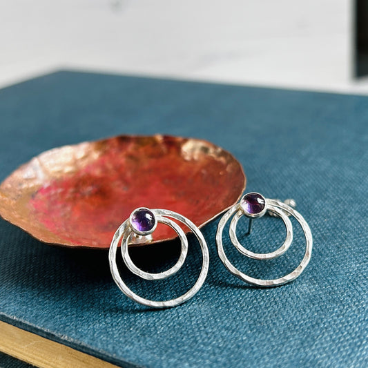 Two small concentric circles handmade in hammered silver make each of these stud earrings, with a small amethyst cabochon at the top of each earring.  Displayed san on a blue book and infront of a copper dish.