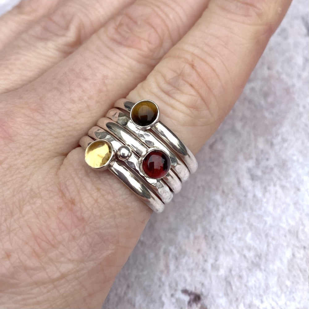 A set of five rings, three smooth slim silver rings each with a semi precious stone set on them, one a Tigers Eye, one a Picture Jasper, and one a Citrine. The other two rings slim textured silver, one with a silver round pebble set on it, by zoe ruth designs