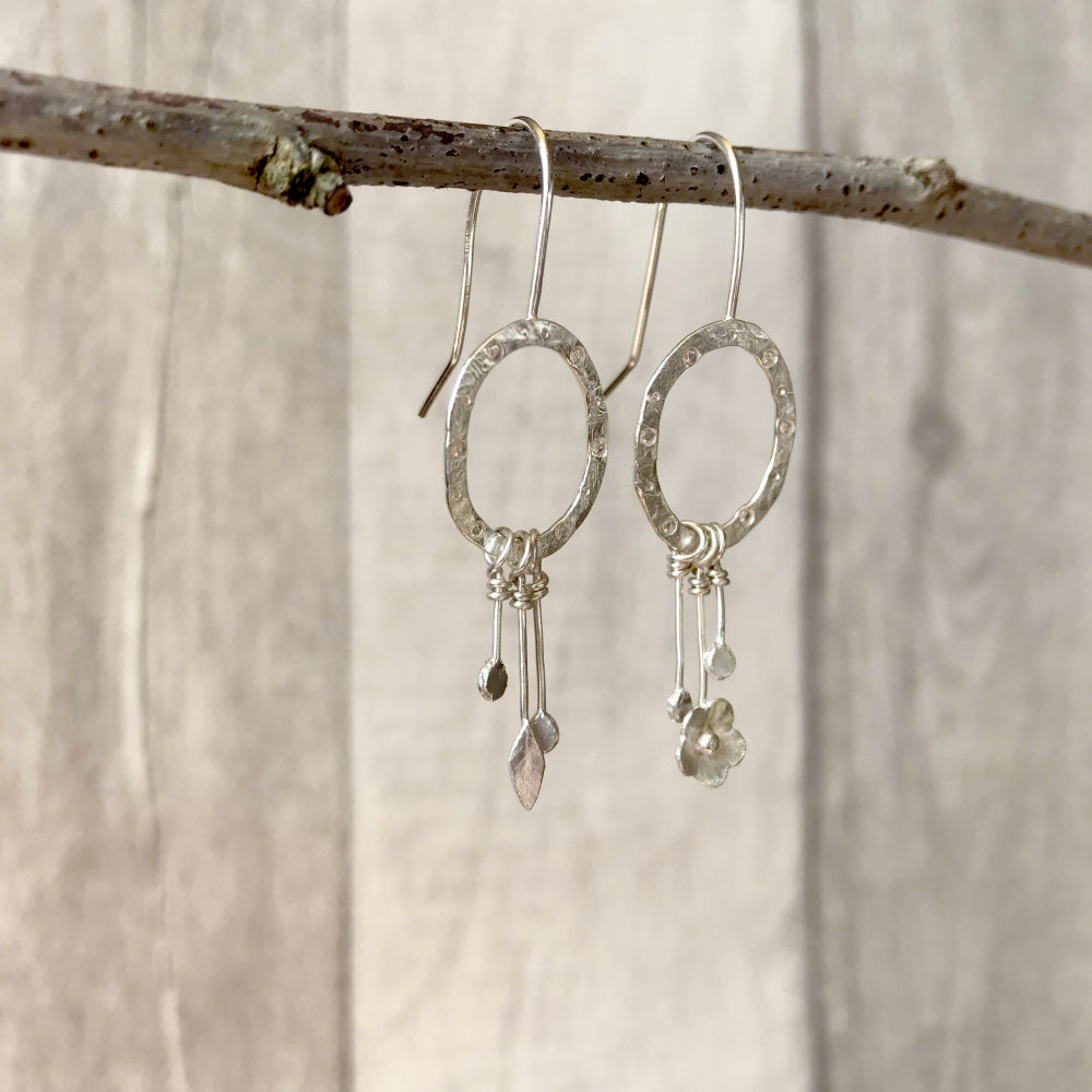 A pair of earrings each a hammered silver oval attached to a hook wire, the oval has a circle and dot pattern stamped on it and hanging as a dangle charm is two bud shapes on each, a tiny leaf on one and a tiny flower on the other. By Zoe Ruth Designs