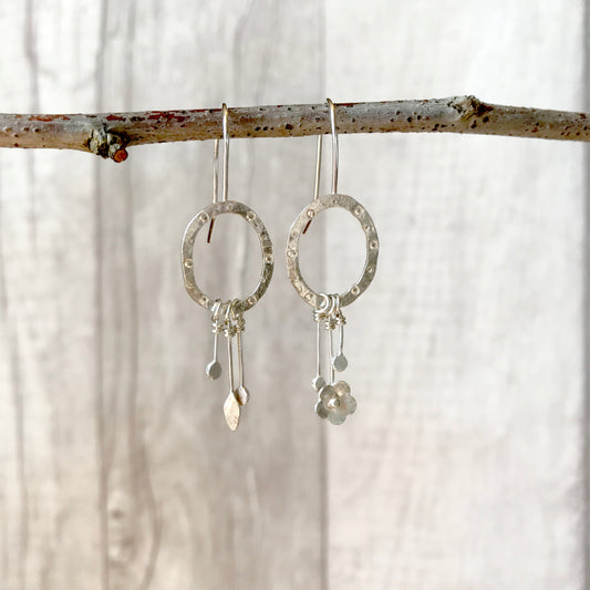 A pair of silver hook style handmade earrings, an oval frame  of hammered silver is soldered to the end of the hook, the oval has a circle and dot stamped pattern.  Each oval has the charm style dangles, on one earring is two drops with a hammered circle and one drop with a tiny handmade silver flower, the other earrings has two drops with a hammered circle and a tiny handmade silver leaf, by zoe ruth designs