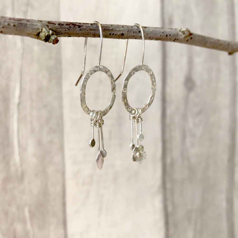 A pair of earrings each a hammered silver oval attached to a hook wire, the oval has a circle and dot pattern stamped on it and hanging as a dangle charm is two bud shapes on each, a tiny leaf on one and a tiny flower on the other. By Zoe Ruth Designs