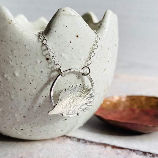 A 2D silver hedgehog with hammered spine texture hangs from some silver trace chain, displayed hung on a ceramic pot, by zoe ruth designs