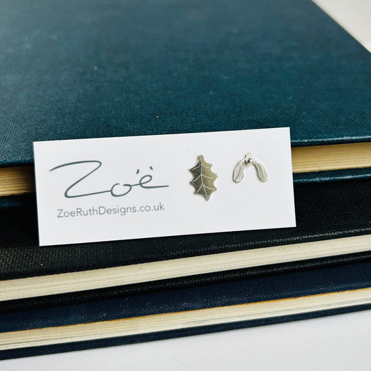 A pair of handmade 2D silver earrings, on a holly leaf with detail scribed one, the other a mistletoe leaf with a silver ball as a berry, displayed on their logo card packaging by zoe ruth designs.