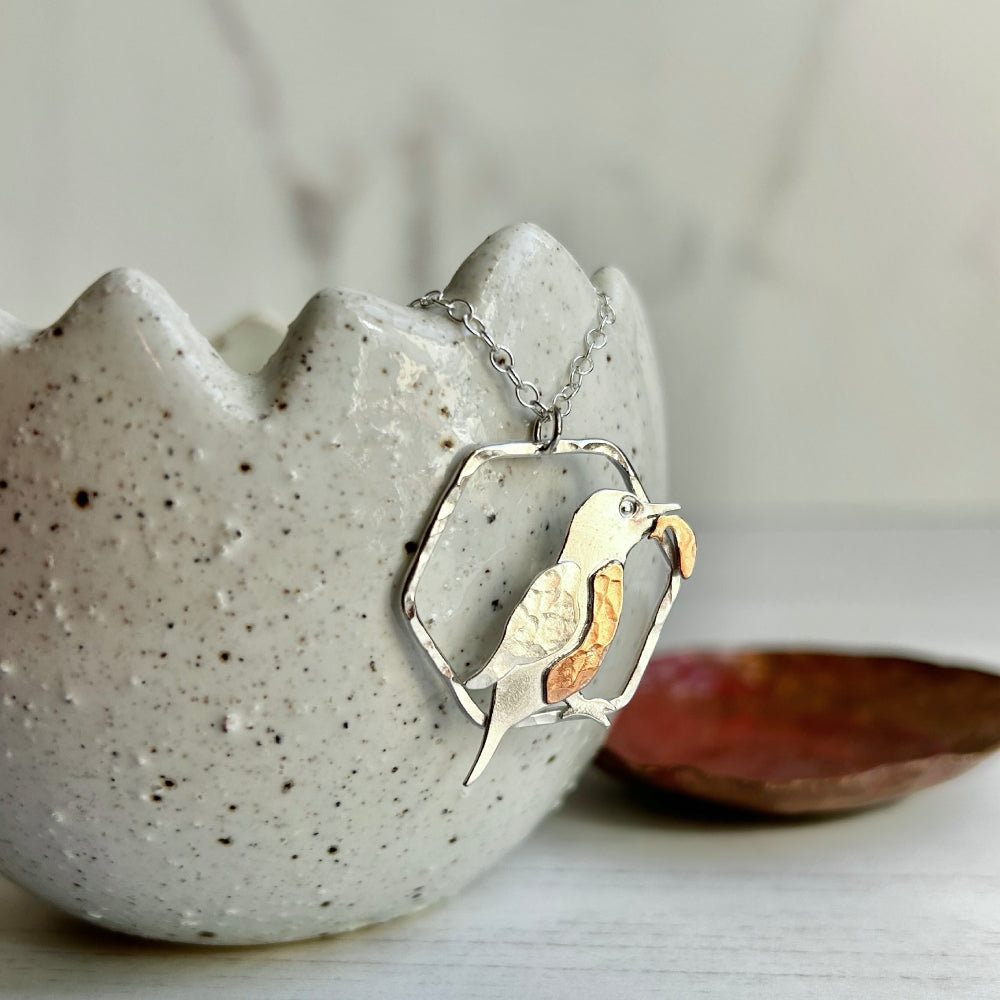 A necklace pendant which is a 2D Kingfisher bird in profile with a fish in its beak, the kingfisher is smooth sterling silver, with a silver textured wing, copper hammered breast and copper hammered fish, displayed hanging over a ceramic pot, by zoe ruth designs