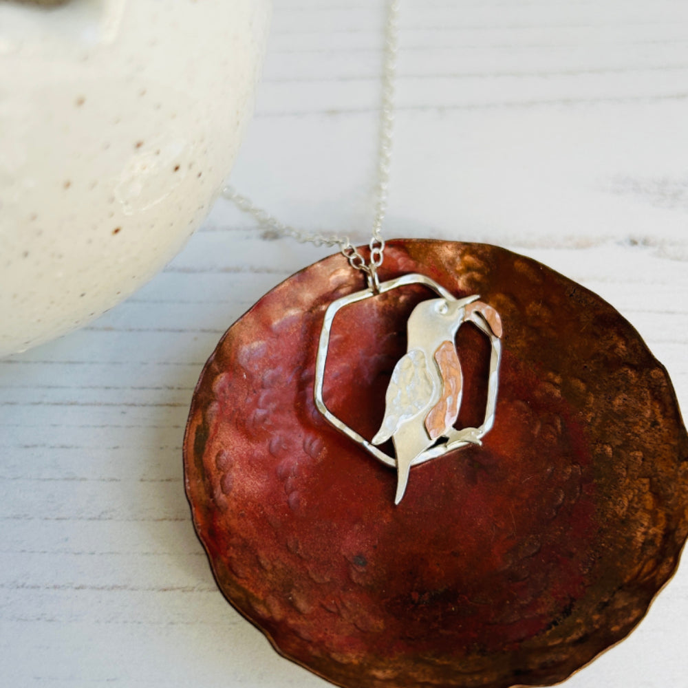A necklace pendant which is a 2D Kingfisher bird in profile with a fish in its beak, the kingfisher is smooth sterling silver, with a silver textured wing, copper hammered breast and copper hammered fish, sat on a copper dish, by zoe ruth designs