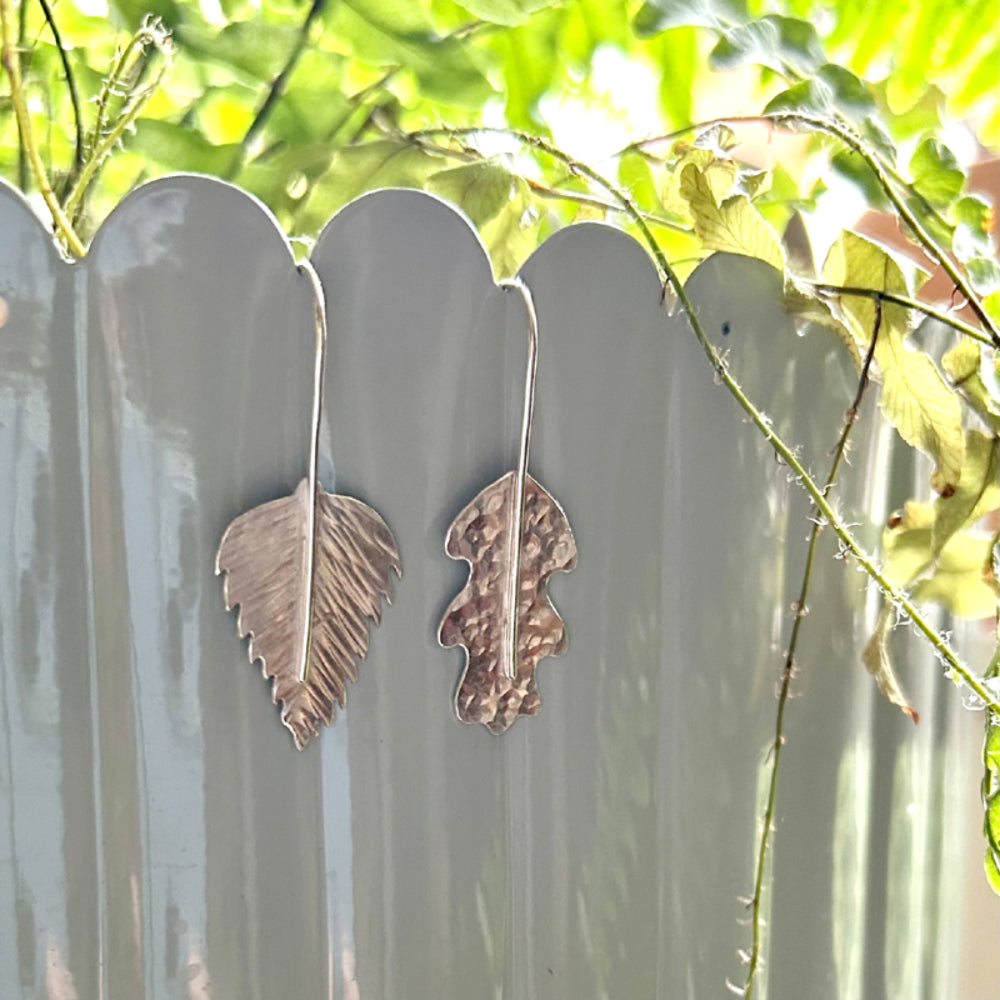 A pair of hand sawn silver leaf earrings, one a dimple hammered oak leaf, the other a silver birch leaf with lines hammered as if leaf vein markings, the silver hooks run down the front of the leaf to be the stem on the designs, by zoe ruth designs