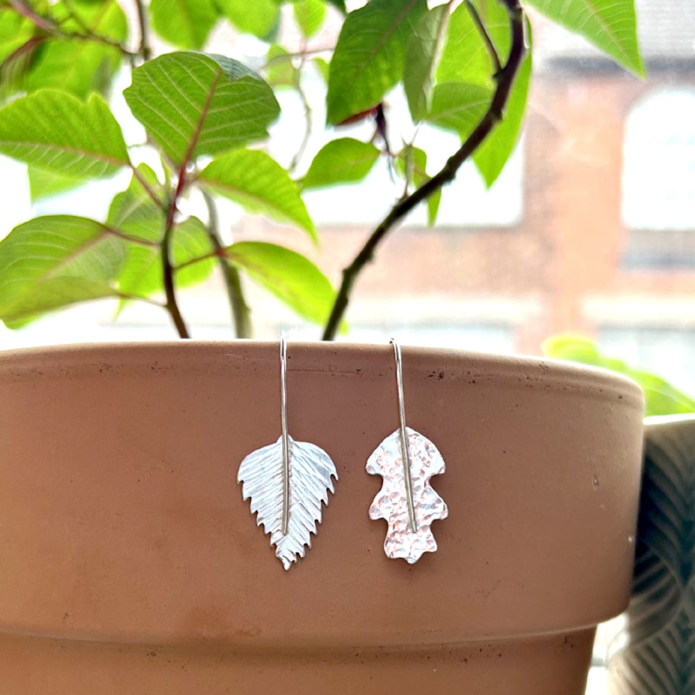 A pair of hand sawn silver leaf earrings, one a dimple hammered oak leaf, the other a silver birch leaf with lines hammered as if leaf vein markings, the silver hooks run down the front of the leaf to be the stem on the designs, by zoe ruth designs