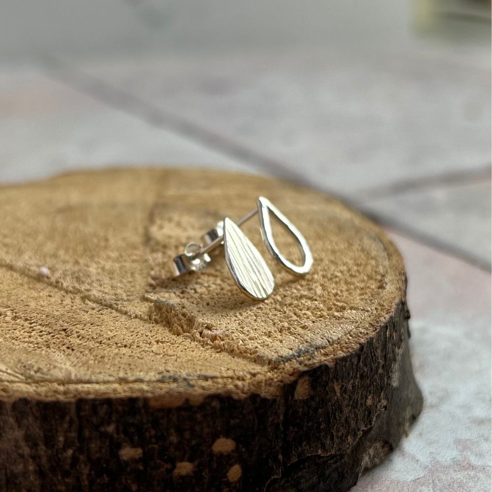 Handmade small silver stud earrings, one a solid teardrop shape with vertical lines texture, the second the outline fo a  teardrop shape with dimple hammered texture.  Displayed sat on sawn log.