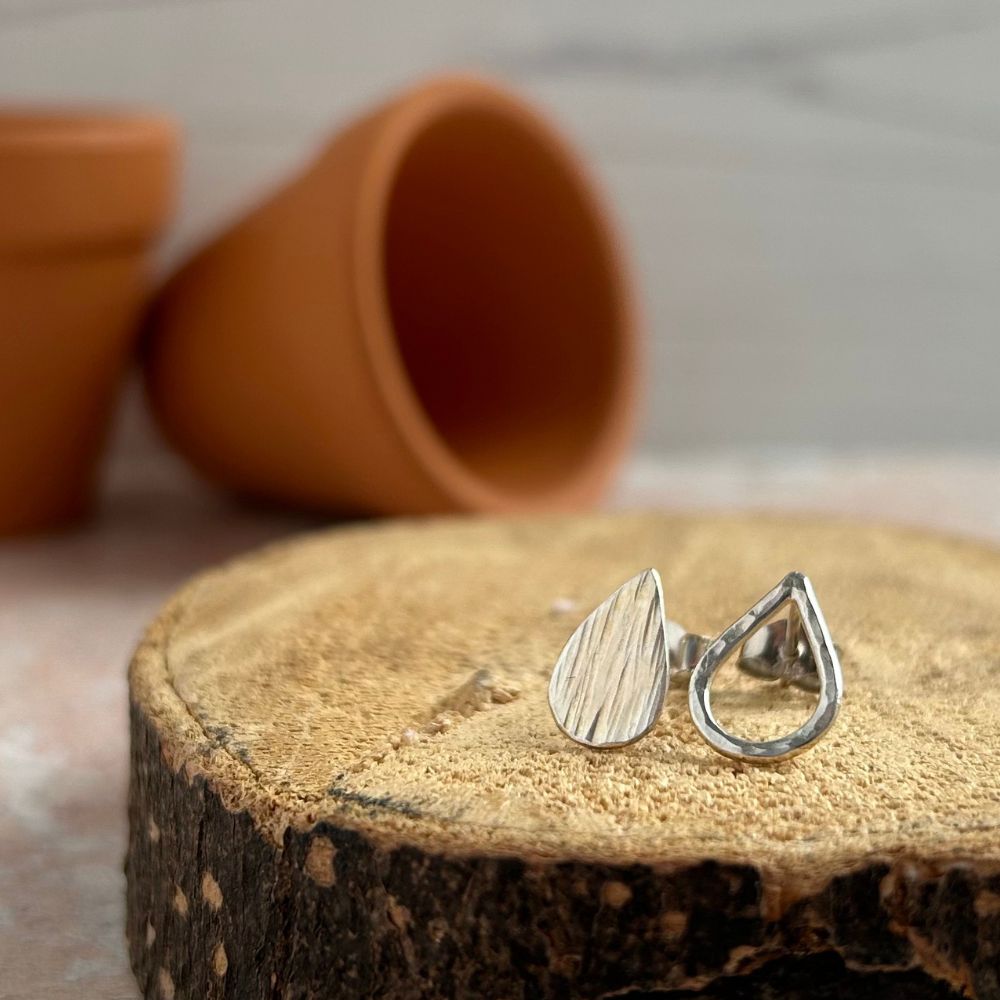 Handmade small silver stud earrings, one a solid teardrop shape with vertical lines texture, the second the outline fo a  teardrop shape with dimple hammered texture.  Displayed sat on sawn log with terracotta plantpots in the background