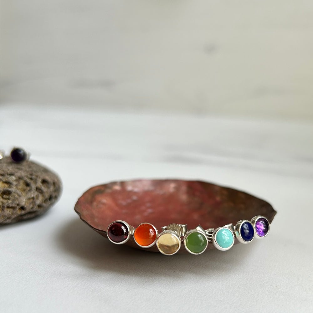 Showing seven colorful gemstone cabochons bezel set in silver to make seven individual stud earrings, displayed on a rustic copper-colored dish with a blurred stone prop in the background.