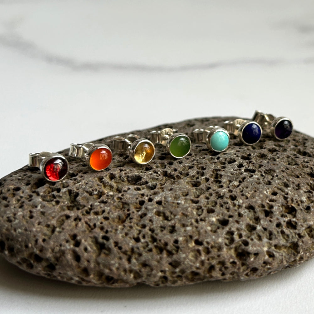 Showing seven colorful gemstone cabochons bezel set in silver to make seven individual stud earrings, displayed on a blurred stone prop.