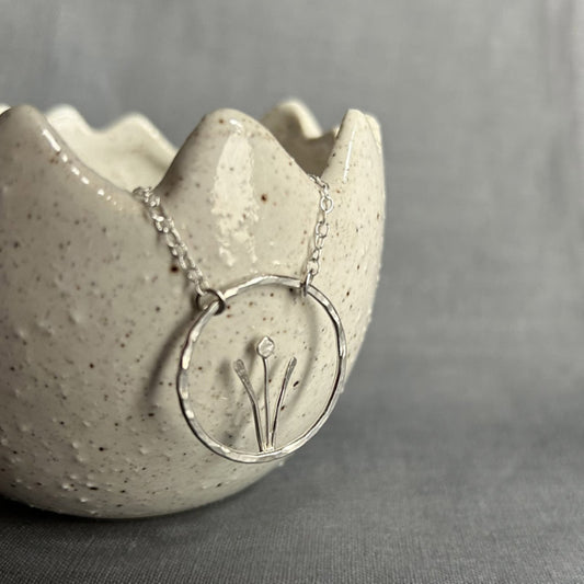 Showing a handmade sterling silver necklace of a spring flower, two tall leaves either side of a stem and bud, framed inside an open silver circle, hung from a silver chain a two points on the circle, by zoe ruth designs