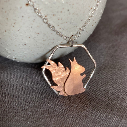 handsawn copper squirrel 2D in profile, smooth body and hammered tail. sat on sterling silver hexagon frame, hanging from a silver chain, by zoe ruth designs