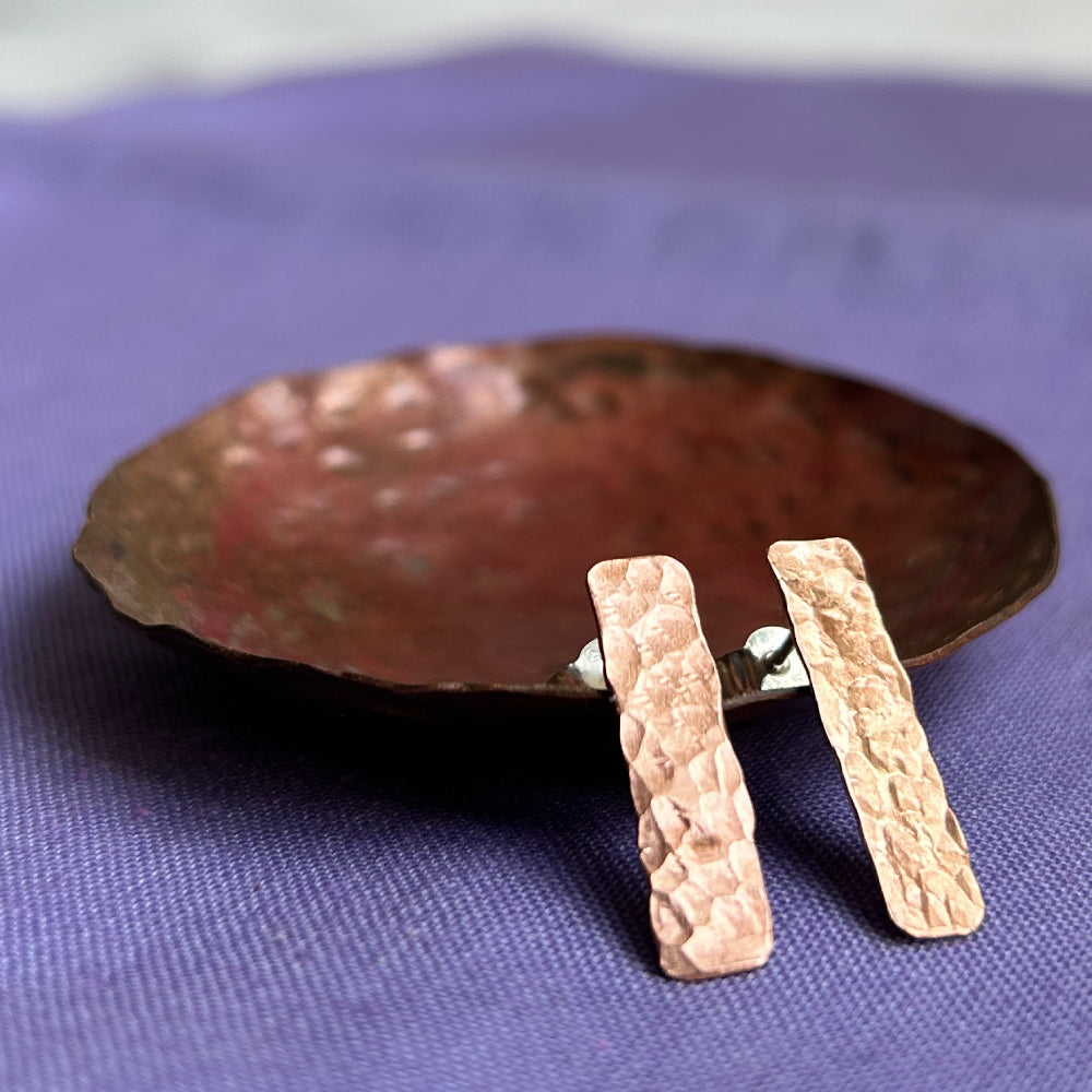 a pair of copper earrings, long rectangle shaped with a hammered surface, displayed on a small dish on a purple cloth