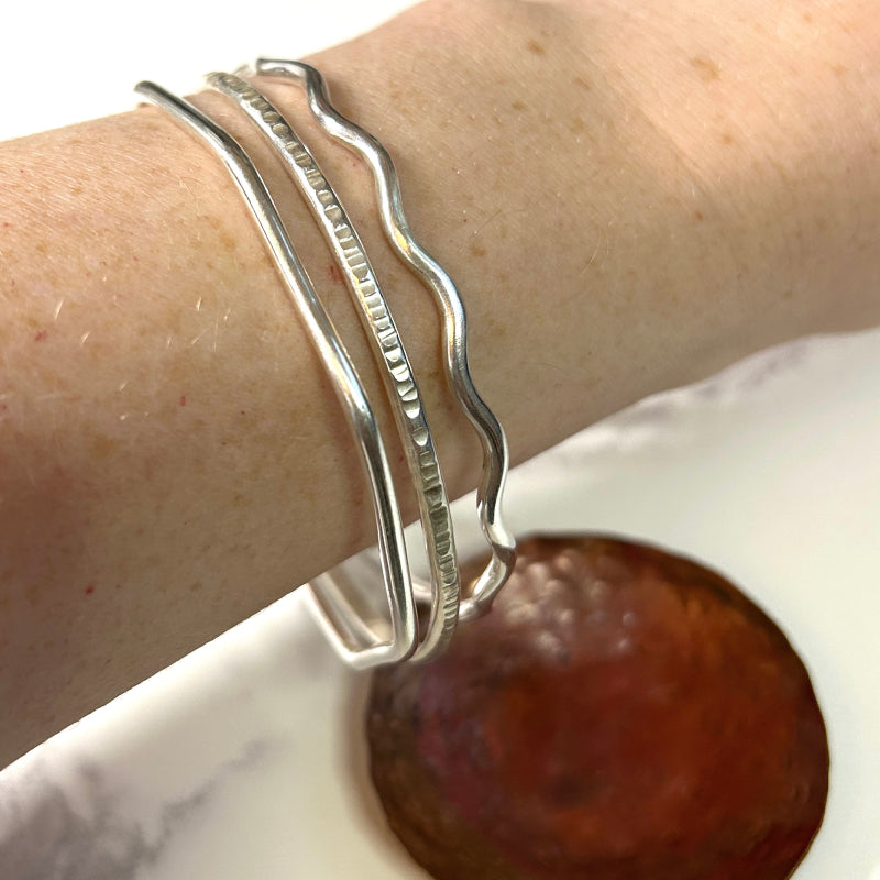 Three different bangles being worn together; a smooth octagon shape, a textured round shape, and a smooth wavy and round shape.