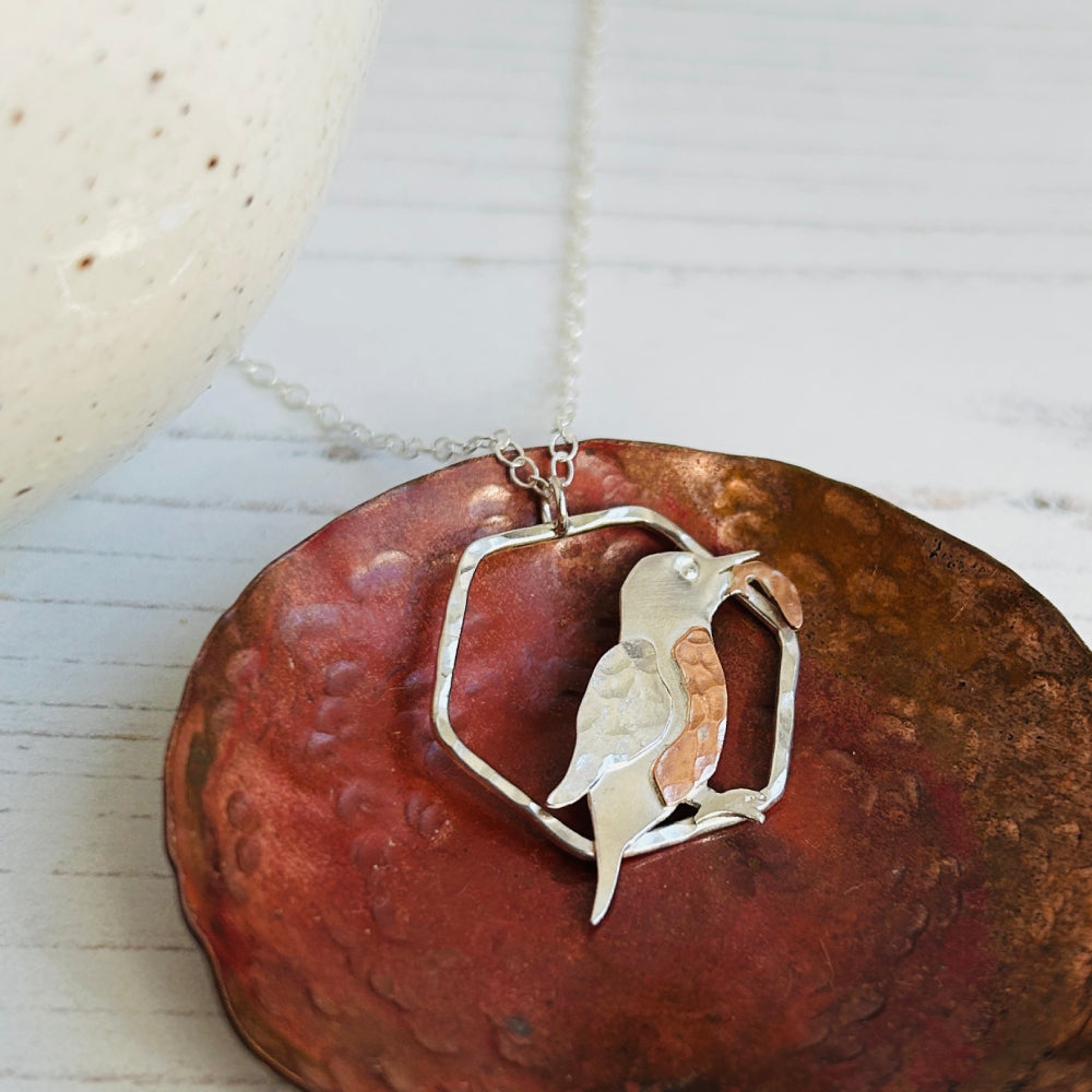 A necklace pendant which is a 2D Kingfisher bird in profile with a fish in its beak, the kingfisher is smooth sterling silver, with a silver textured wing, copper hammered breast and copper hammered fish, sat on a copper dish, by zoe ruth designs