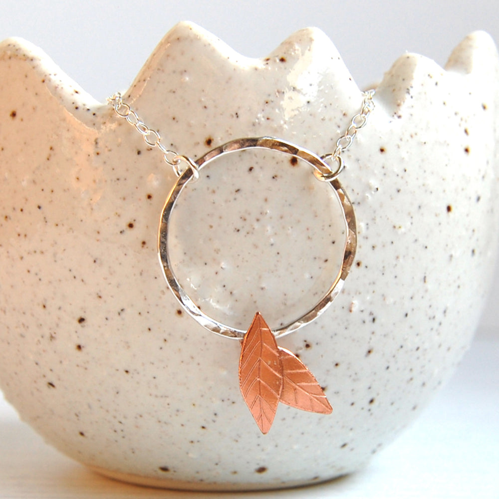 Showing a close up front view of a handmade necklace, it is a silver open circle with two copper leaves fixed at the bottom, by zoe ruth designs