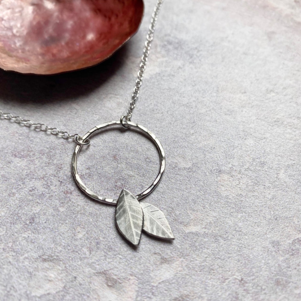 A handmade silver pendant of an open silver circle with two 2D silver leaves attached if falling off of the bottom, by zoe ruth designs 