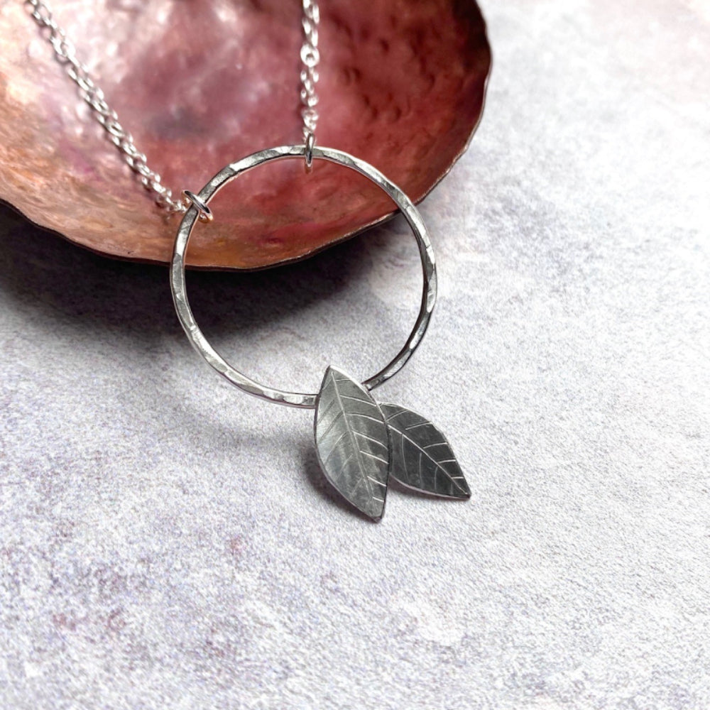 A close up of a handmade silver pendant of an open silver circle with two 2D silver leaves attached if falling off of the bottom, by zoe ruth designs 