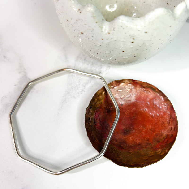 Sterling silver octagon shaped bangle, laying down next to a round copper dish to see the shape