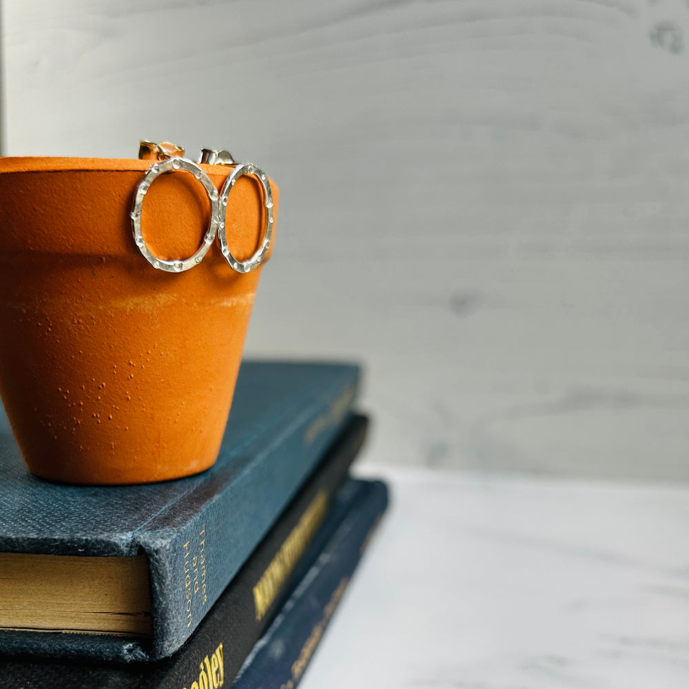 a pair of handmade silver stud earrings, oval in shape with a circle and dot stamped texture by zoe ruth designs. styled on a ceramic pot on a stack of books