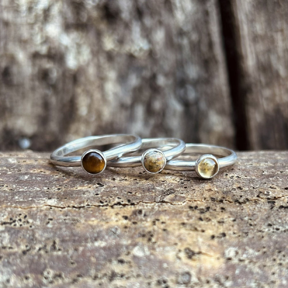 Three seperate cabochon set stones  on silver rings are displayed ontop of a wooden surface, there is a Tigers Eye stone, Picture Jasper stone and a Citrine stone