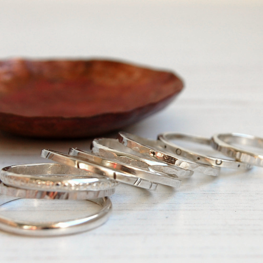 Showing nine silver stacking rings scattered next to a copper dish all with different textures, by zoe ruth designs.