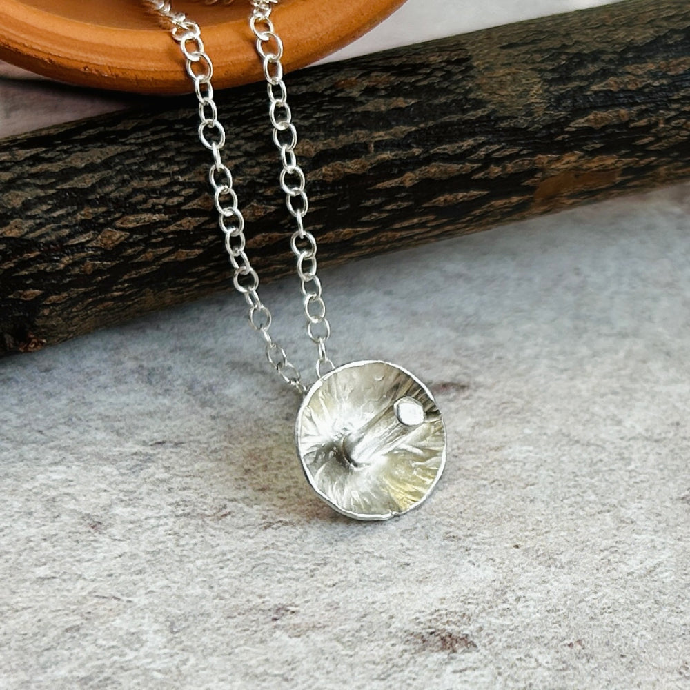 A silver chain necklace with a textured, handmade silver toadstool pendant showing the underside texture, resting on a piece of wood on a light gray stone surface.
