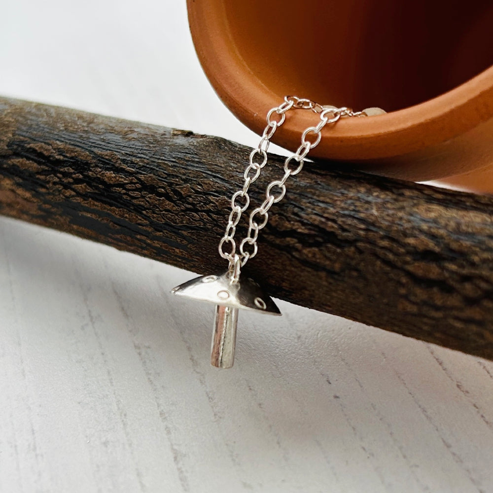 A silver chain necklace with a small mushroom-shaped pendant, styled on a textured gray surface with a dark wood branch and a terracotta plant pot in the background.