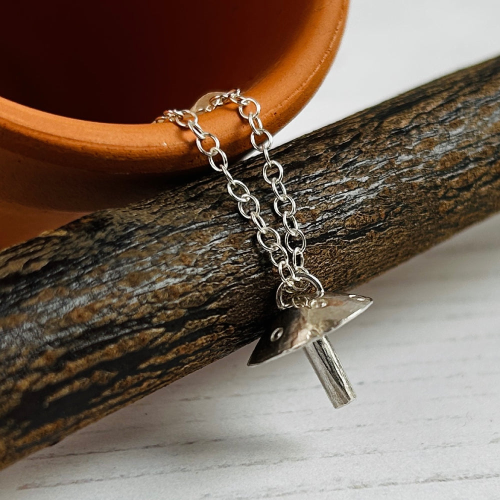 A silver chain necklace with a small mushroom-shaped pendant, draped over a dark textured branch with part of a terracotta plantpot visible in the background.