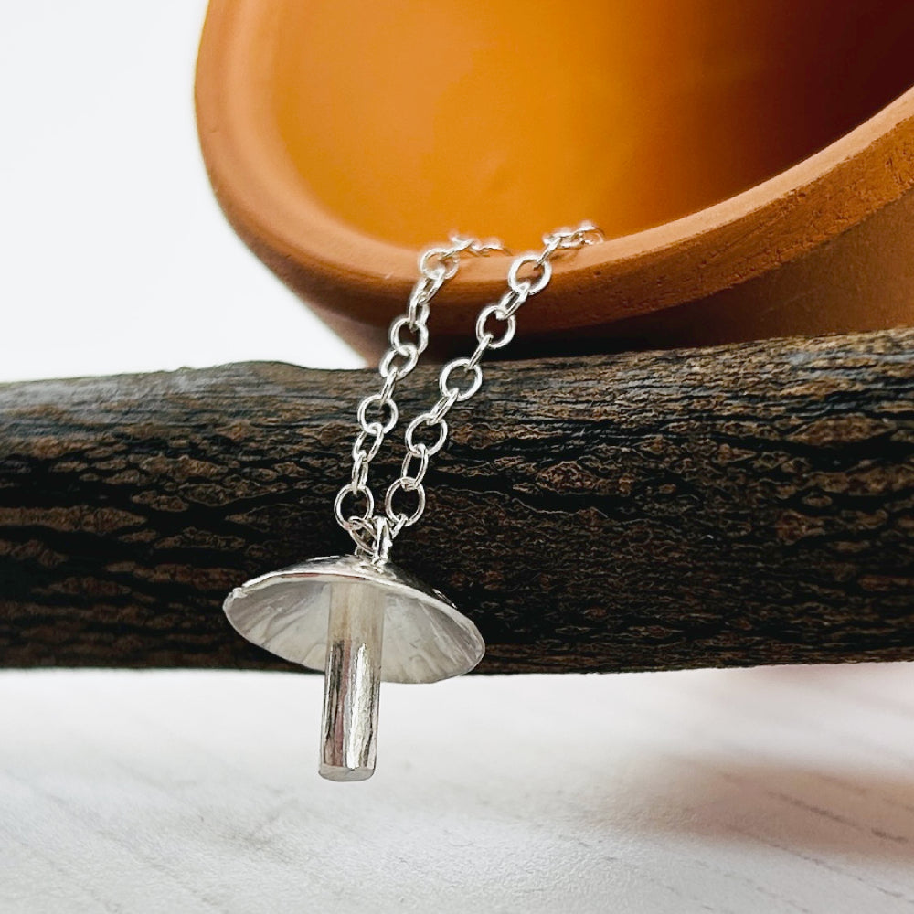 A silver mushroom-shaped pendant necklace on a delicate silver chain, draped over a dark textured branch with a terracotta pot in the background.
